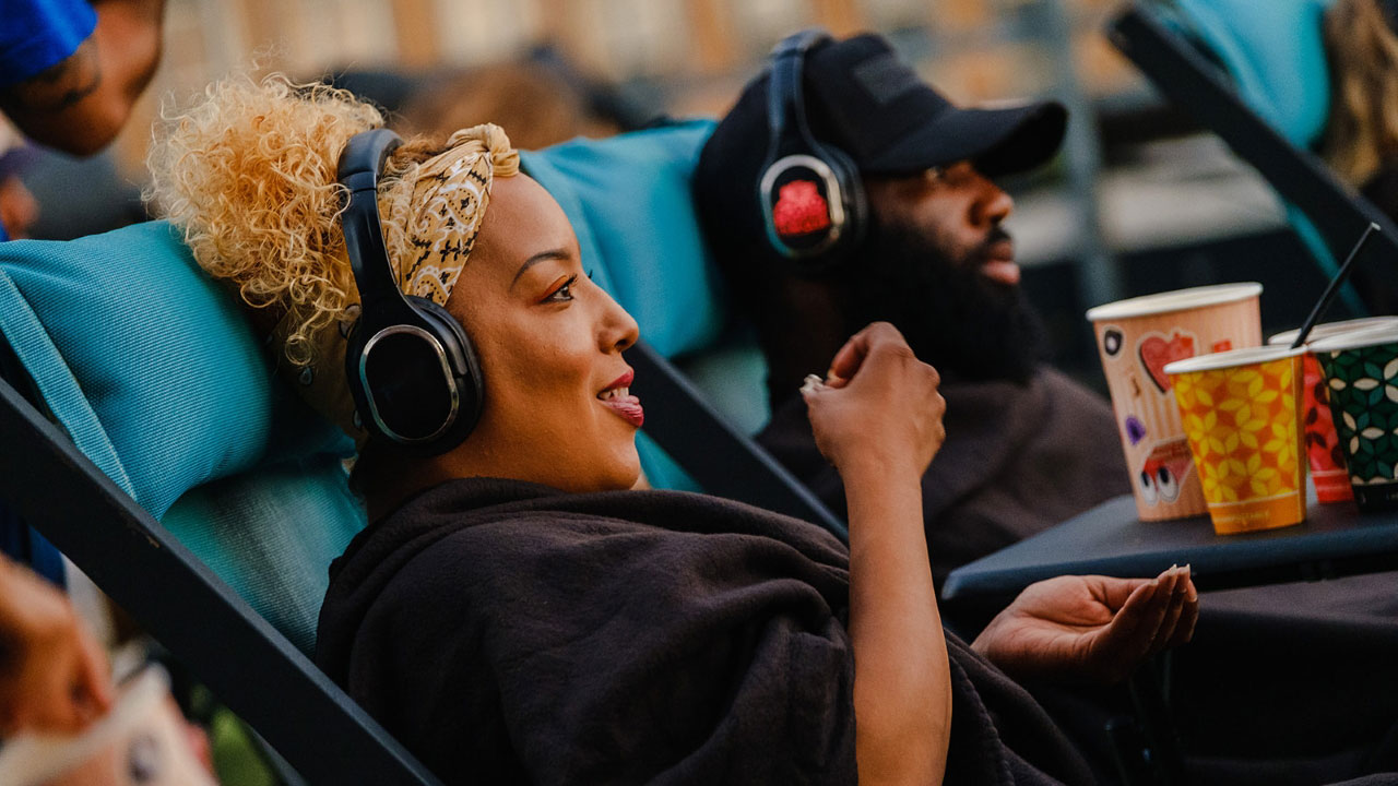 A photo of a man and woman sat in comfy chairs next to each other at Rooftop Film Club Stratford smiling at the screen with headphones on while eating snacks