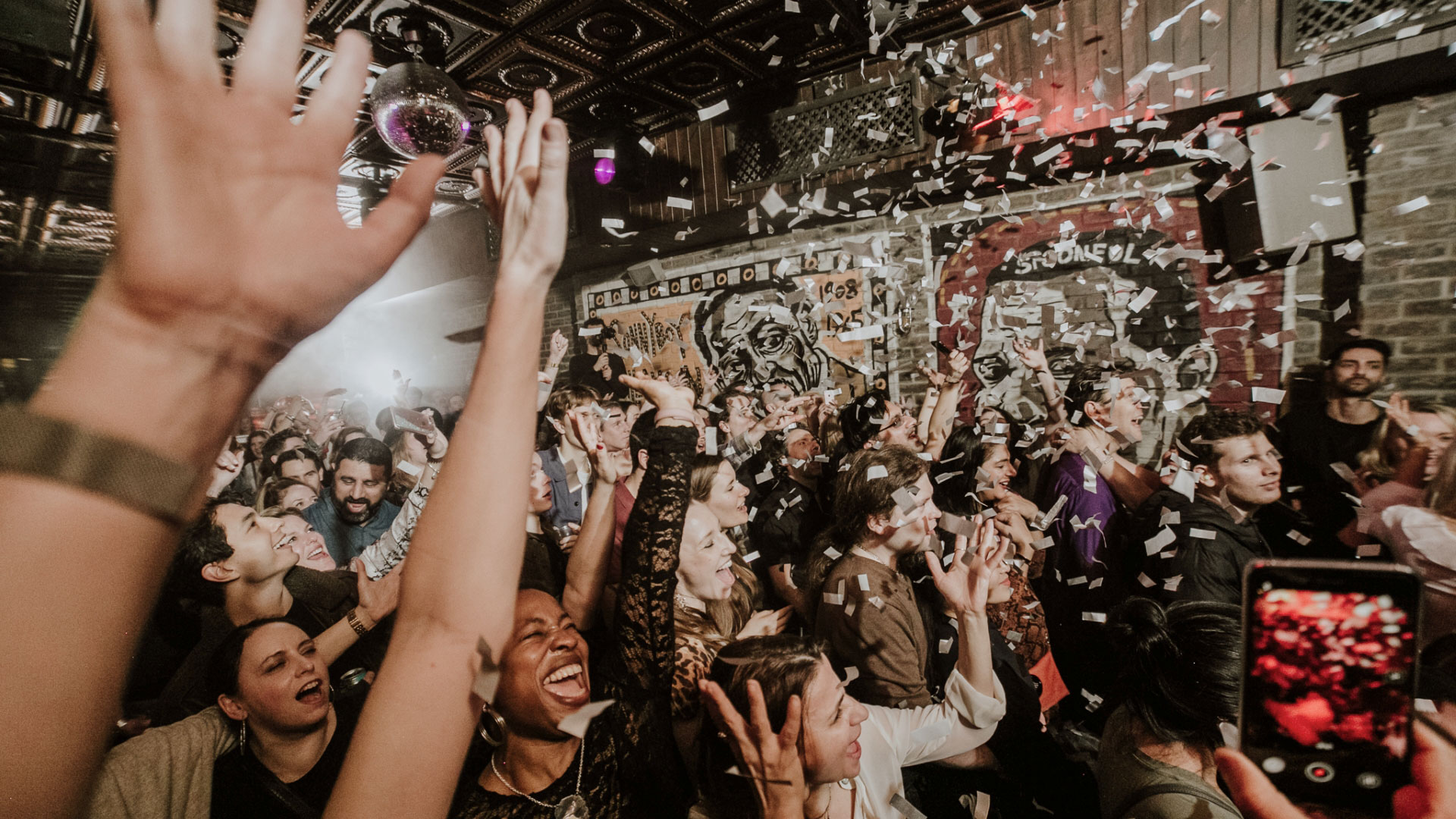 A photo of the crowd at Blues Kitchen in Camden. Everyone is smiling with their hands in the air facing the stage as confetti falls from above