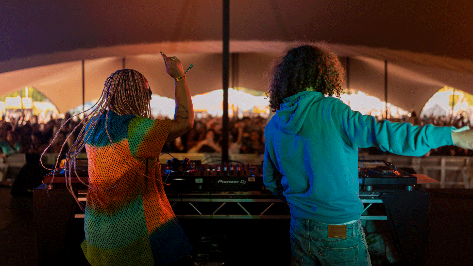 A girl in a rainbow shirt and man in a blue hoodie DJ in a tent in front of large audience