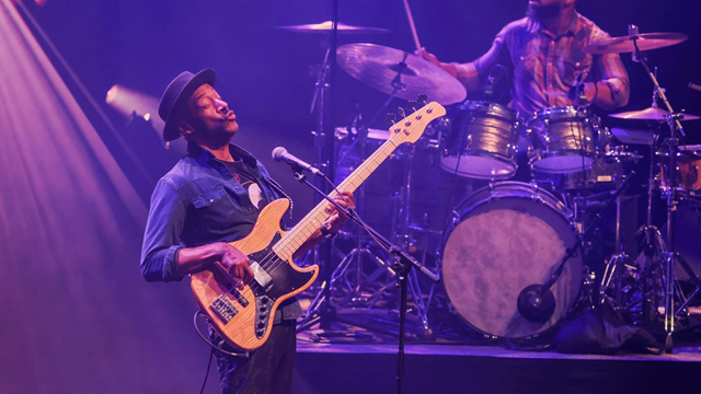 Marcus Miller plays the guitar in a blue lit room with drummer in the background