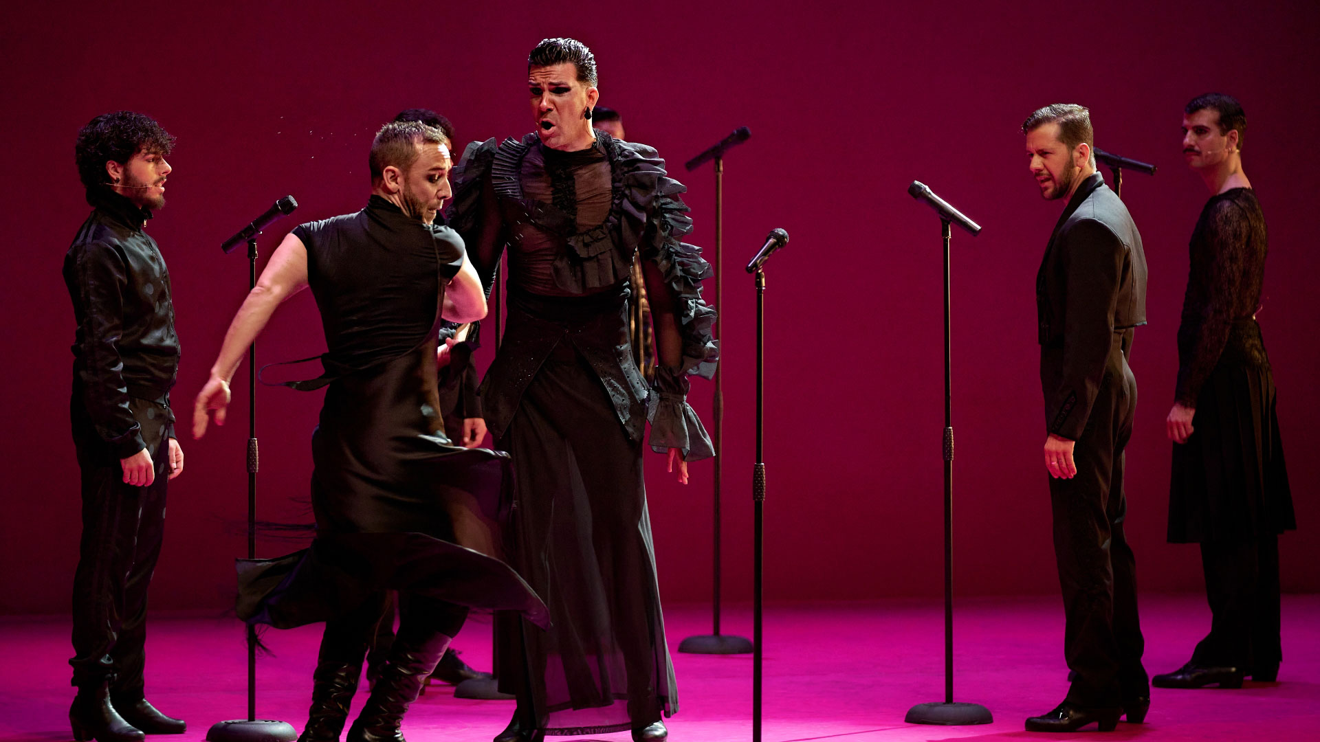 A photo of five men dressed in black on stage with microphones dancing and singing with a pink background