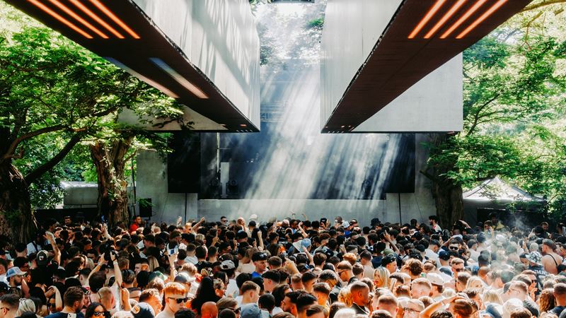 See Barking Park transformed into an otherworldly landscape of sound, performance and art installations at High Lights Festival. Credit: Jake Davis. Image courtesy of Khroma Collective. People dancing underneath trees and concrete structure during a daytime festival performance.