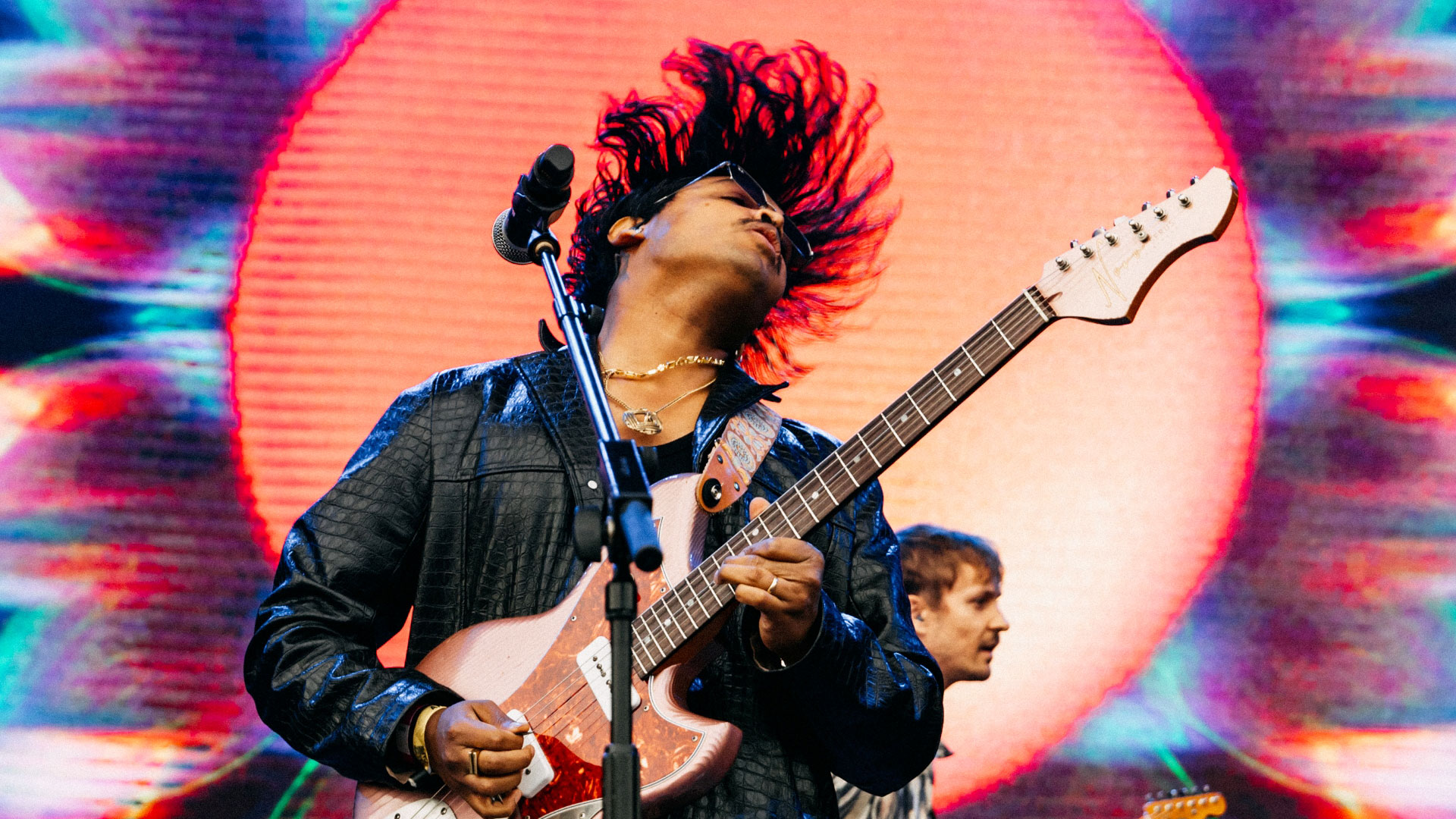 A close up photo of a musician playing a guitar whilst throwing his head back with a microphone in front of him and psychedelic visuals on the screen behind