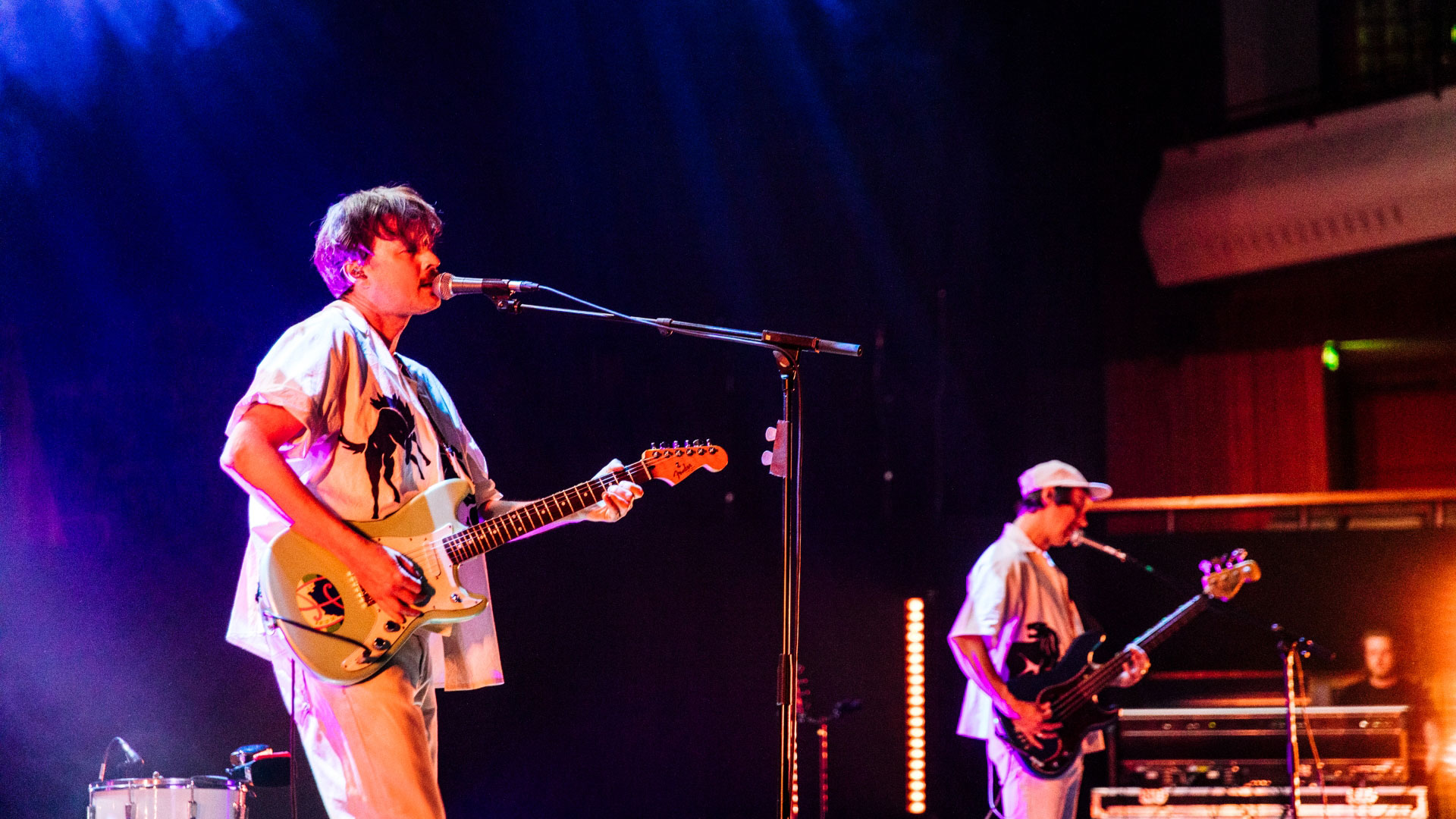A photo of two men performing onstage each with a guitar whilst singing into a microphone and colourful lights behind them 