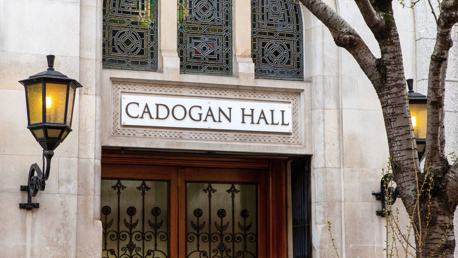 A photo of the exterior of Cadogan Hall with the venue name placard and lamps on the building with a tree in the foreground
