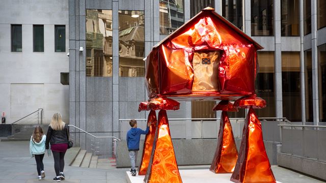 Orange marble sculpture of a house on stilts surrounded by grey buildings.