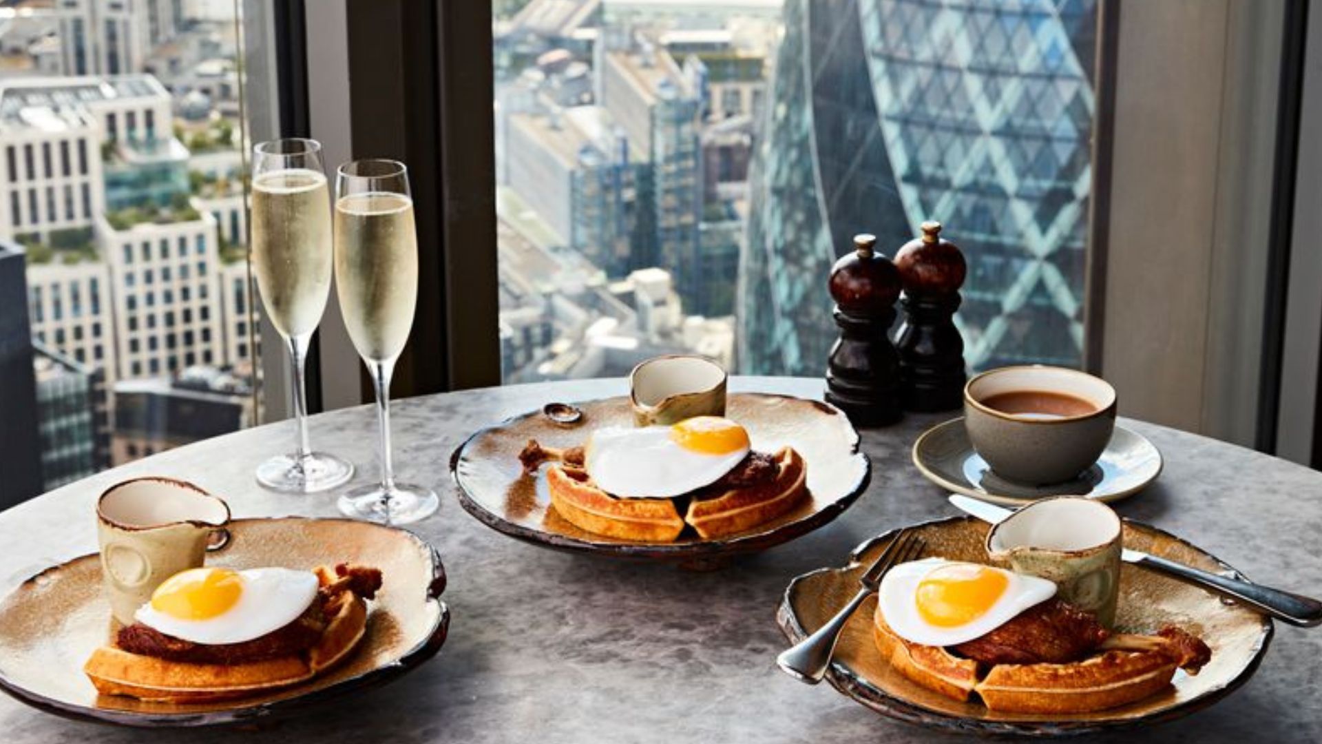 a grey marble round table with 3 plates of golden eggs and duck leg on waffles, cups of tea, champagne flutes and the Gherkin just out of focus in the background