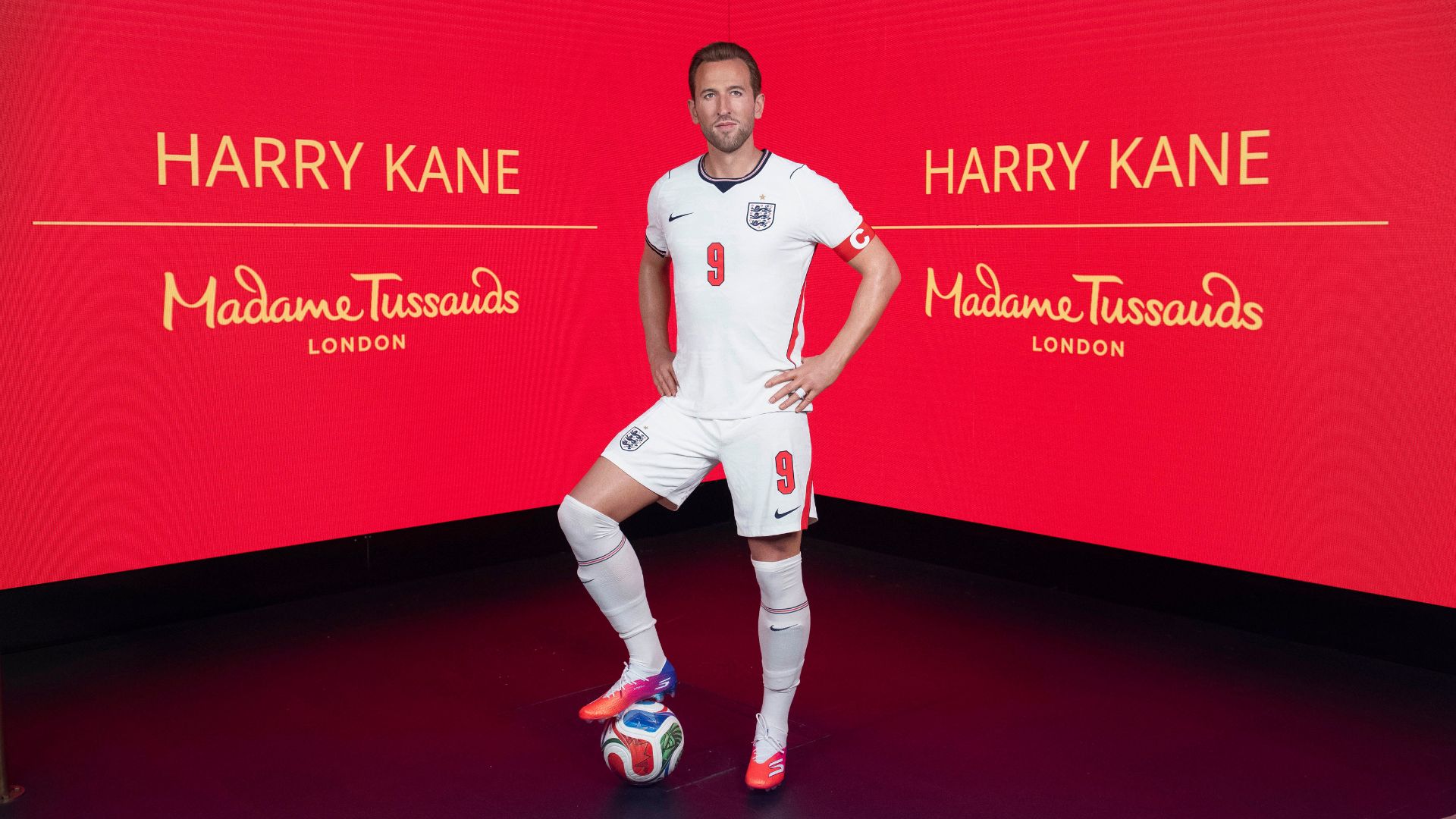 Wax figure of Harry Kane in full football kit with red Madame Tussauds screen behind.