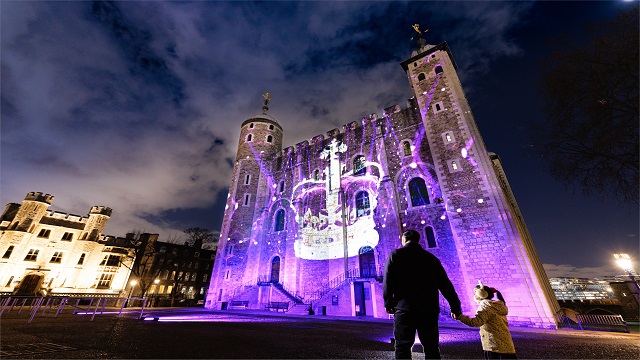 Low angle shot of the Tower of London, with a light show projected onto the building and the silhouette of an adult and a child holding hands watching the display