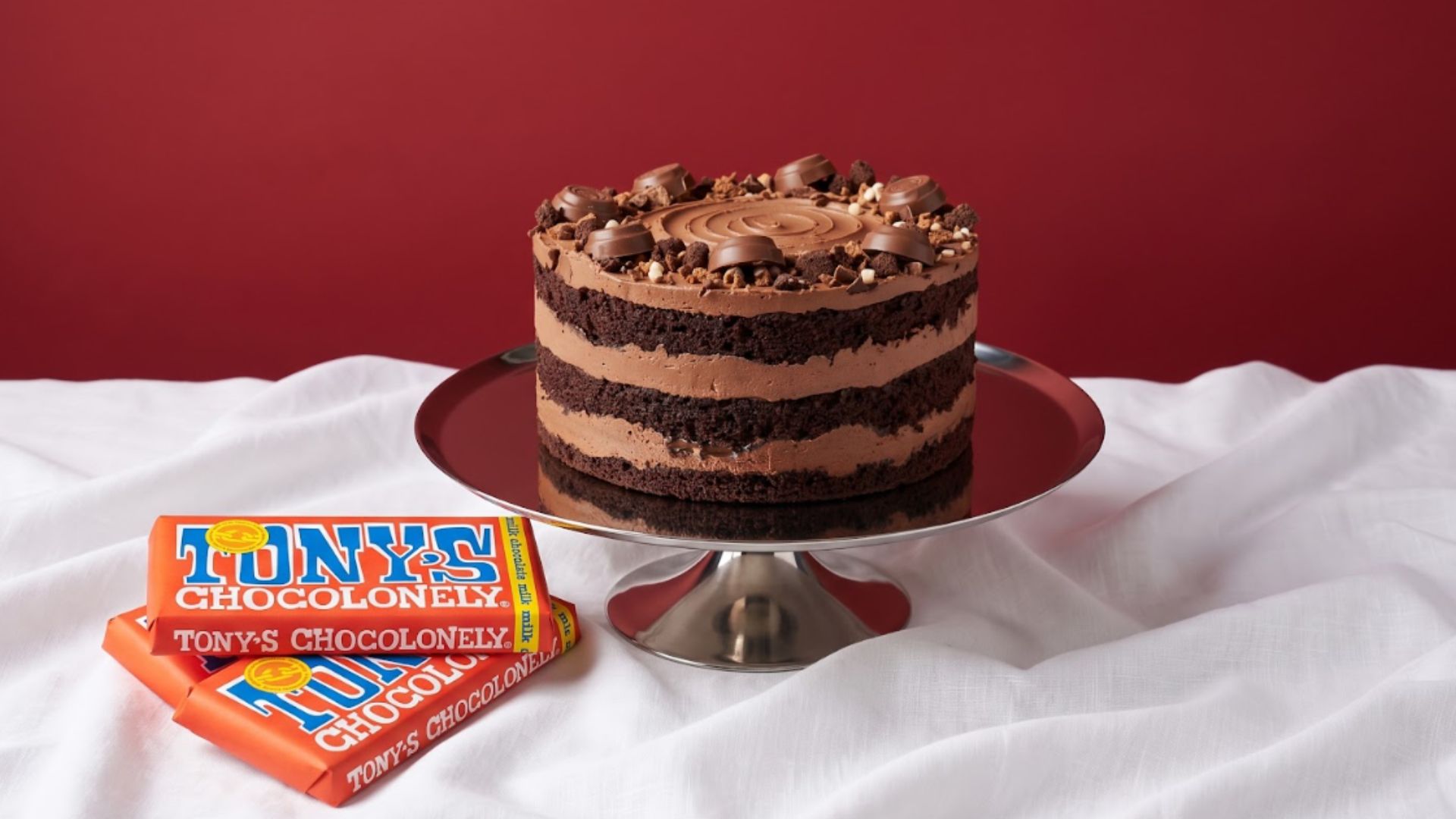 Chocolate layer cake topped with chocolate pieces on a silver cake stand, with Tony's Chocolonely chocolate bars in the foreground.