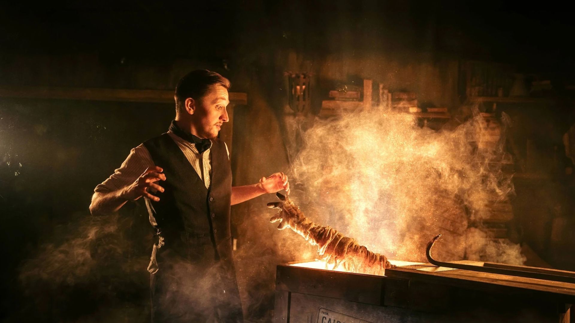 Performer beside a glowing crate as a wrapped mummy's arm emerges through smoke during The London Dungeon's Curse of the Mummy experience.