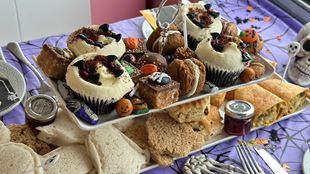 A Halloween-themed afternoon tea spread featuring cupcakes decorated with black bats and orange pumpkins, assorted pastries, scones, finger sandwiches, and small jars of jam on a two-tier stand. The table is covered with a purple spiderweb-patterned cloth and scattered Halloween decorations, including miniature pumpkins and skeleton cutlery.