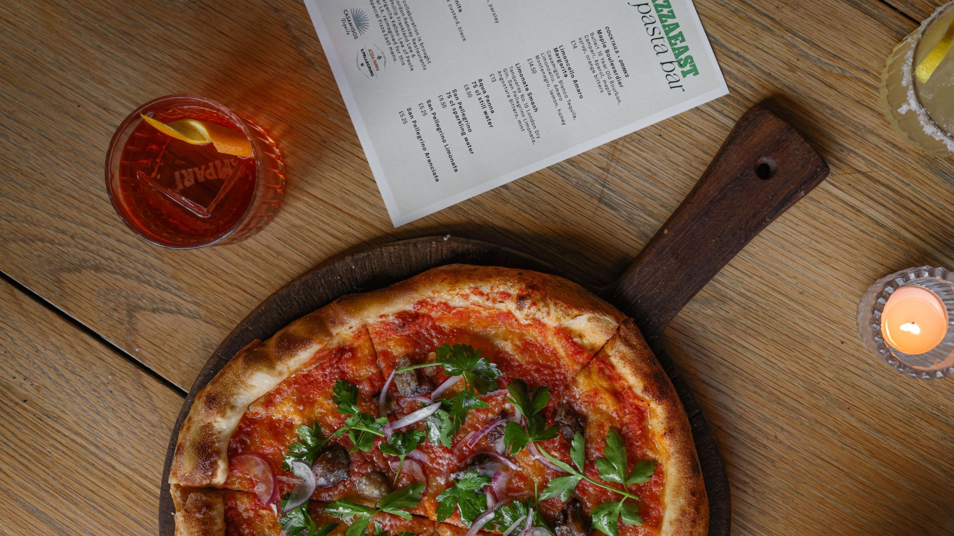 A circular pizza in the foreground on a table with a menu for a pasta bar pop-up in the background.