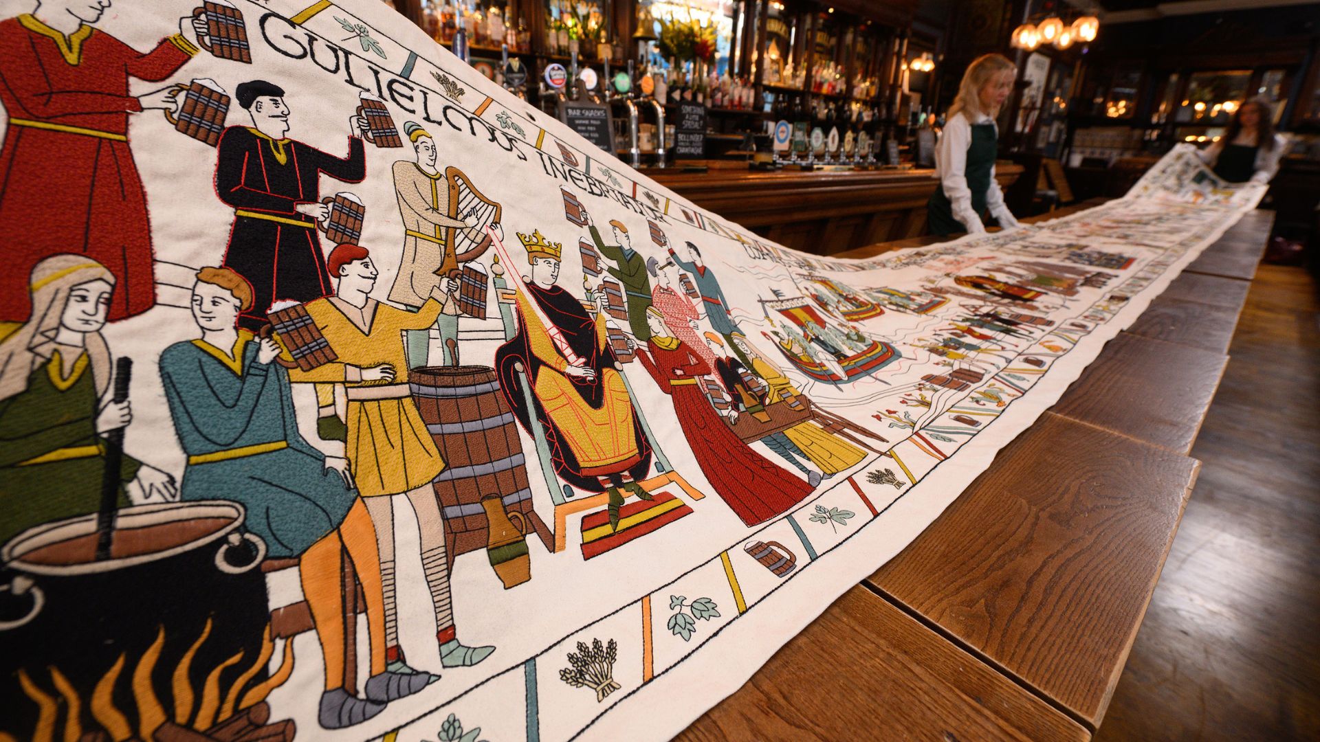 a long wooden table in a pub, with a long, embroidered tapestry detailing the history of beer, held by people in suits and white gloves