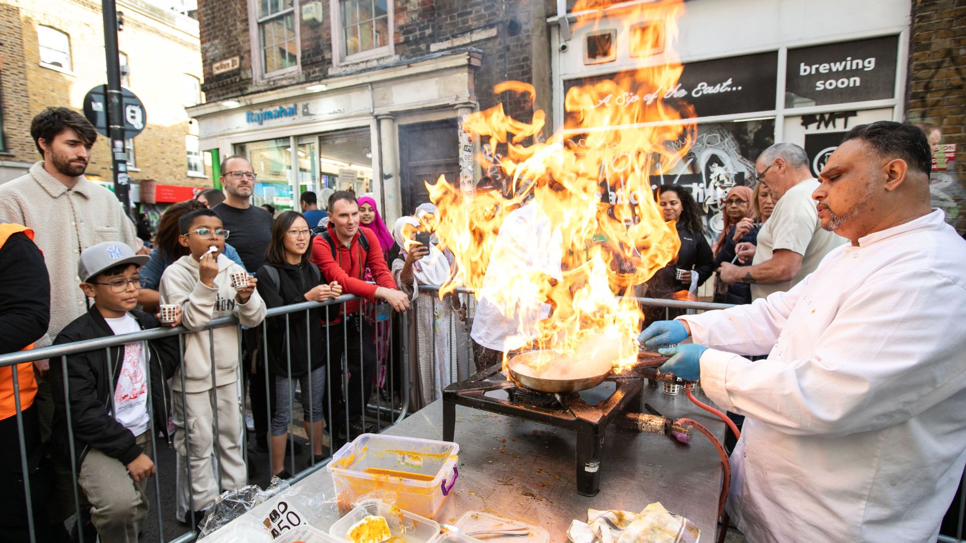 a chef set up on a cooking table outside on a street, holding a pan with flames, whilst of smile and watch from behind a barrier