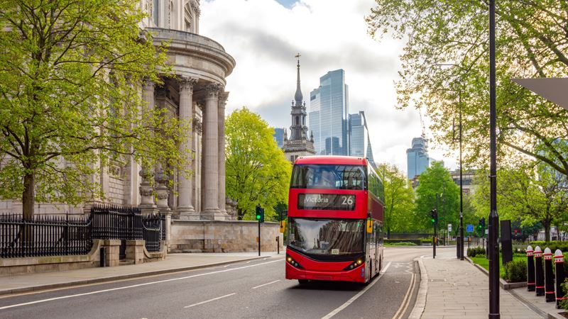 Navigate London for a small cost using the vast London bus network. Credit: Shutterstock. Image courtesy of Shutterstock. A red double decker bus drives along a road in London with a stately building and modern offices in the background and leafy trees lining the road