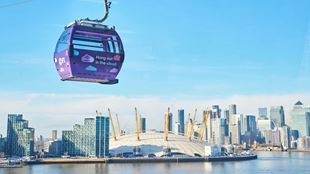 A photo of the O2 Arena and Canary Wharf high-rise buildings with the river Thames in the foreground and a London Cable Car hanging overhead