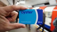 A London transport Oyster card. Image courtesy of Shutterstock. Person holding a blue London transport Oyster card with a London Underground train arriving at a Tube platform in the background.