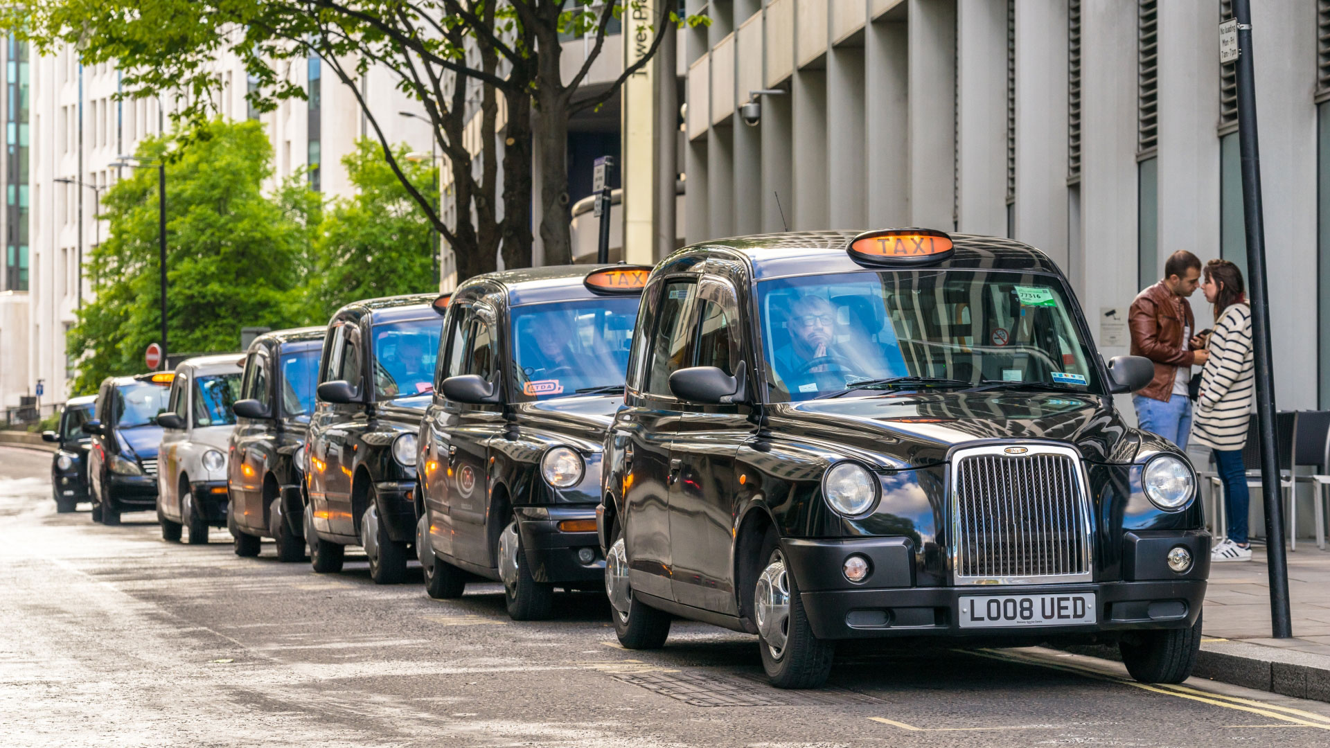 A photo of a row of black London taxi cabs lining up at the side of a road waiting for passengers
