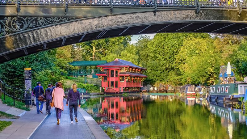A photo of the back of two women walking along Regent's Canal with a floating restaurant and houseboats and a bridge in the foreground