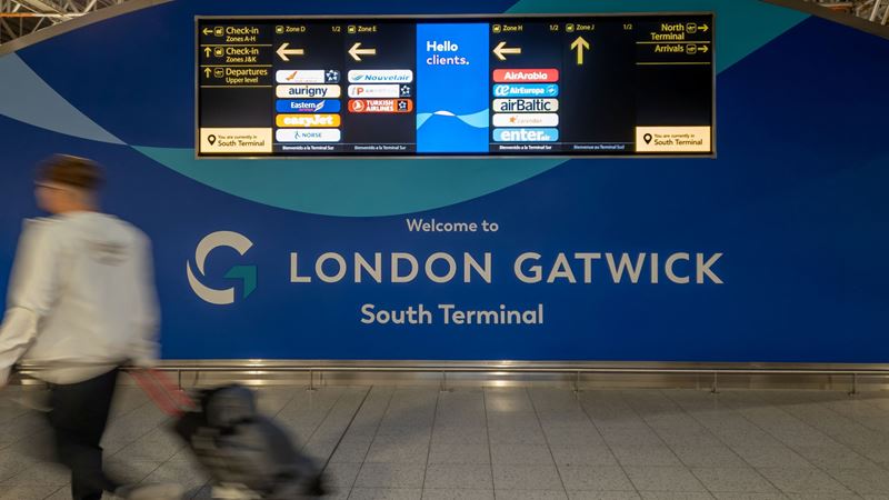 A photo of a man with a suitcase walking past a Welcome to London Gatwick south terminal sign with digital signs showing flight information