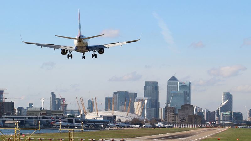 Find the cheapest ways to travel to and from London. Credit: Shutterstock. Image courtesy of Shutterstock. A photo of an airplane landing on the runway at London City Airport with the City of London skyline in the background