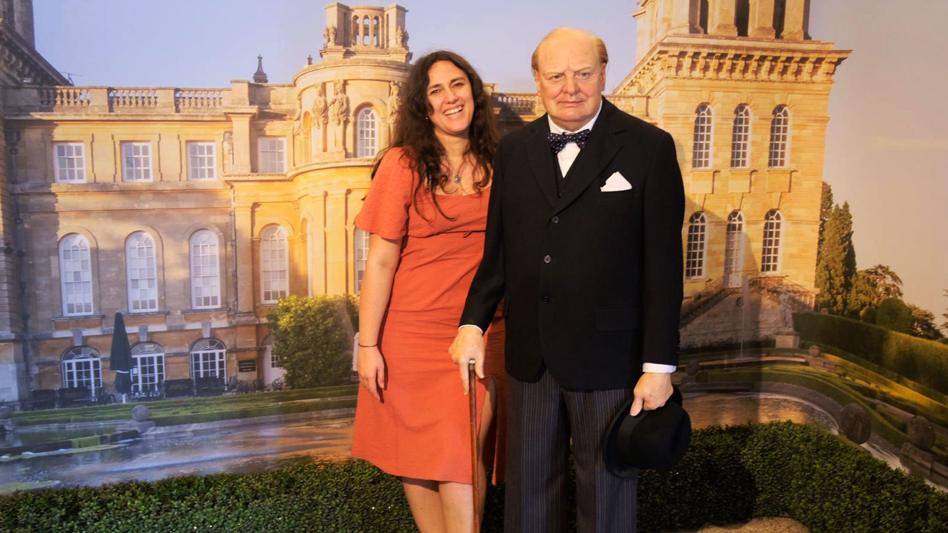A woman in a rose dress stands next to a wax figure of Winston Churchill with Bleinheim Palace in the background.