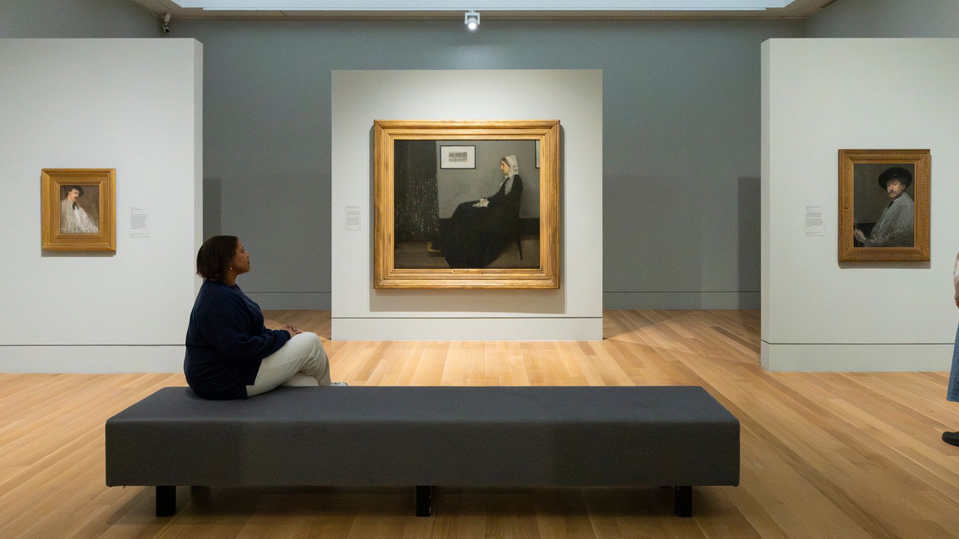 Paintings on wall with Portrait of His Mother in the centre and a person sitting down facing it.