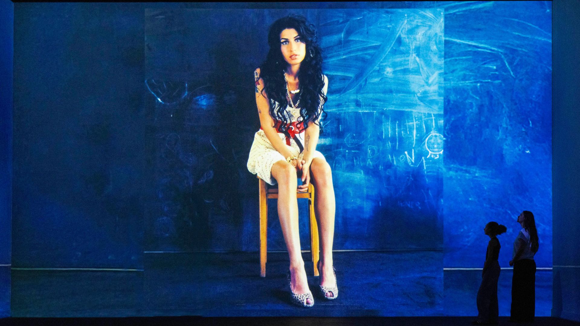 Large-scale portrait of Amy Winehouse on a blue background at FRAMELESS with two people looking up at it.