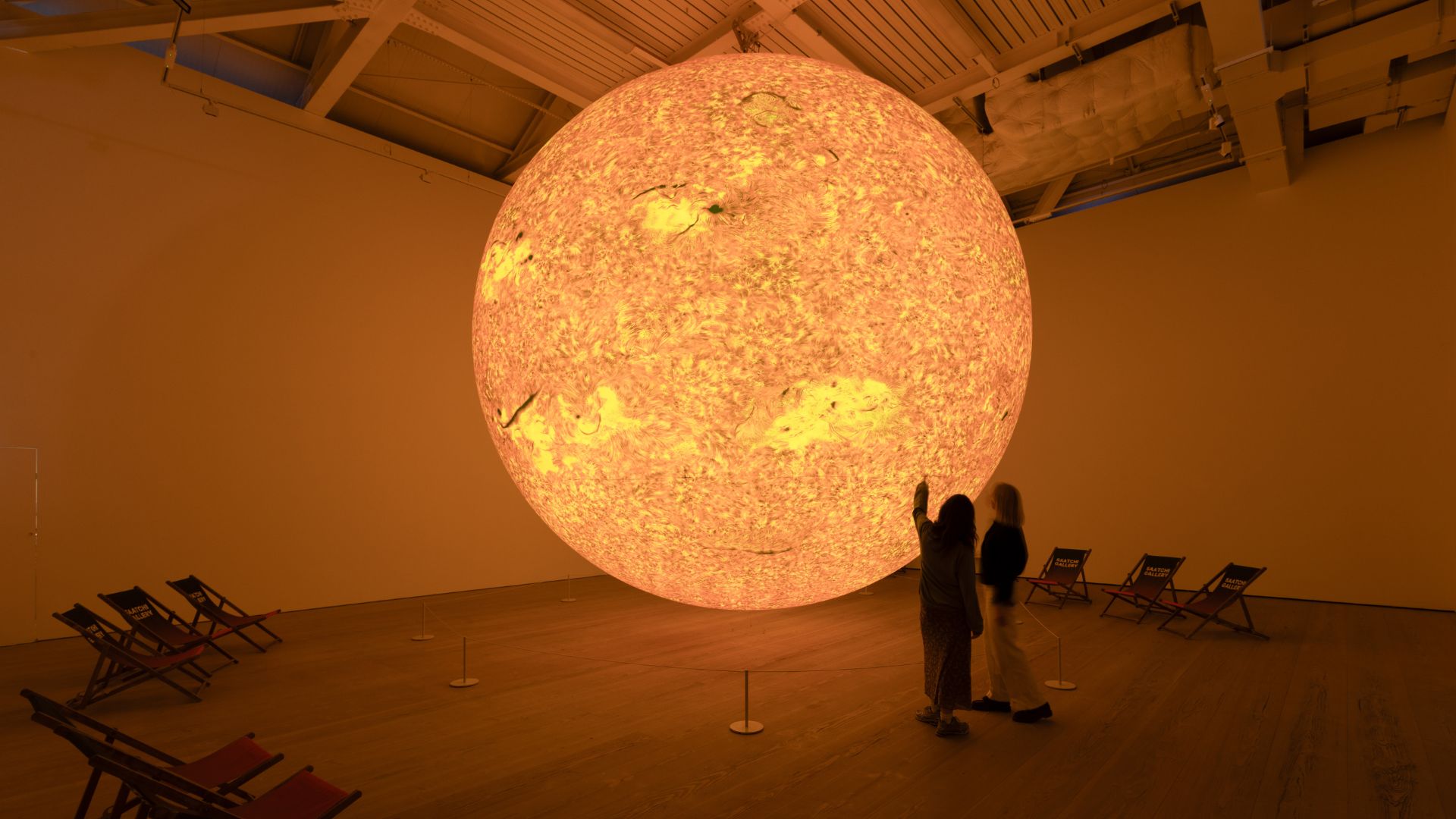 Large-scale sculpture of the sun with two people standing next to it in gallery room.