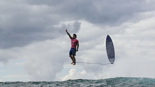 Photo of Gabriel Medina holding up one arm with his surfboard behind him during the Paris 2024 Olympic Games.