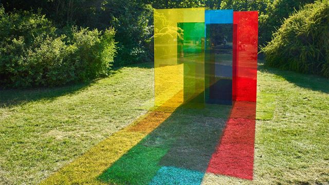 Four vertical pieces of coloured glass (yellow, green, blue and red) of varying heights, cast coloured shadows on the grass