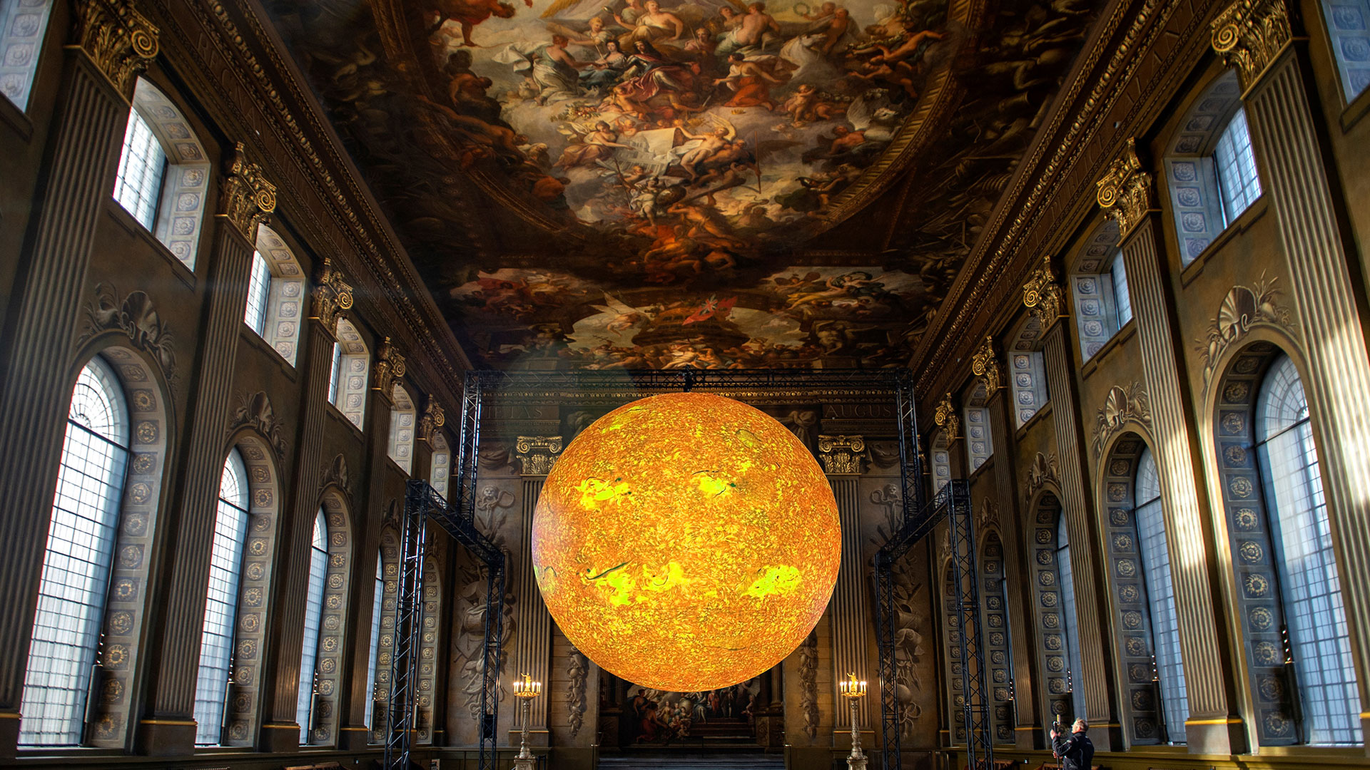 Admirez la nouvelle installation époustouflante de Luke Jerram au Painted Hall, où une magnifique structure représentant le soleil flotte gracieusement dans ce cadre historique. Avec l'aimable autorisation de l'Old Royal Naval College. Dans le Painted Hall, un bâtiment grandiose avec un plafond peint de style Renaissance, un globe jaune lumineux est suspendu dans l'air, symbolisant le soleil.