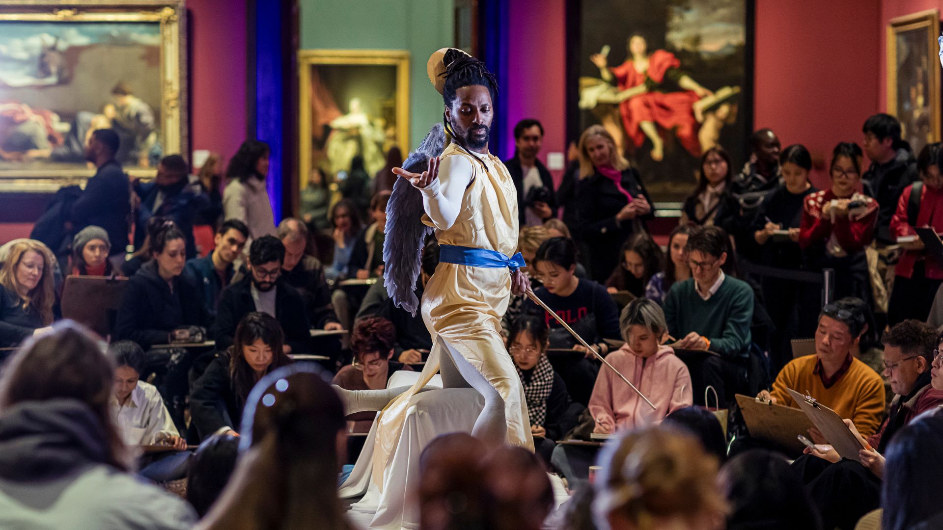 Life drawing session with model standing in the centre of gallery room and multiple people gathered around.
