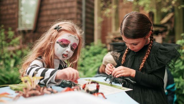 Two children in Halloween costumes take part in a spooky craft activity outdoors.
