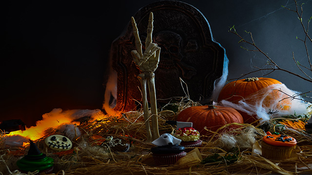 Ein quietschbunter Arm ragt aus dem Boden in den Himmel, auf dem mehrere Kürbisse und Halloween-Muffins verteilt sind.