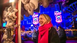 Une jeune femme blonde portant une écharpe rouge se trouve devant un stand où sont suspendus des ours en peluche.