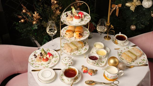 Festive afternoon tea setting with a three-tier stand holding assorted pastries, scones and finger sandwiches on floral china plates. The table includes cups of tea, wine glasses, bowls of clotted cream and jam, and decorative Christmas ornaments, set against a backdrop of holiday greenery and gold decorations.