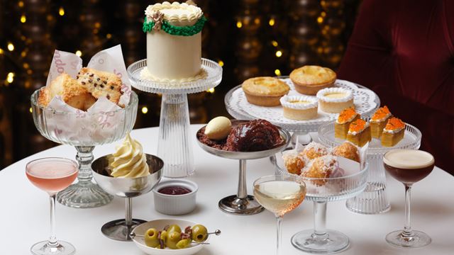 Festive dessert and cocktail spread featuring a small decorated cake on a glass stand, assorted pastries including mince pies and sponge squares, a bowl of scones, and various drinks such as cocktails and champagne, all arranged on a white table against a backdrop of warm golden lights.