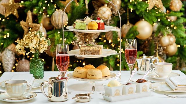 Festlicher Nachmittagstee mit einer dreistöckigen Etagere mit verschiedenen Backwaren, Sandwiches und Scones, dazu zwei Gläser Rosé-Sekt. Der Tisch ist mit edlem Porzellan, einer silbernen Teekanne und Gewürzen gedeckt, im Hintergrund steht ein geschmückter Weihnachtsbaum mit goldenen Ornamenten und funkelnden Lichtern.