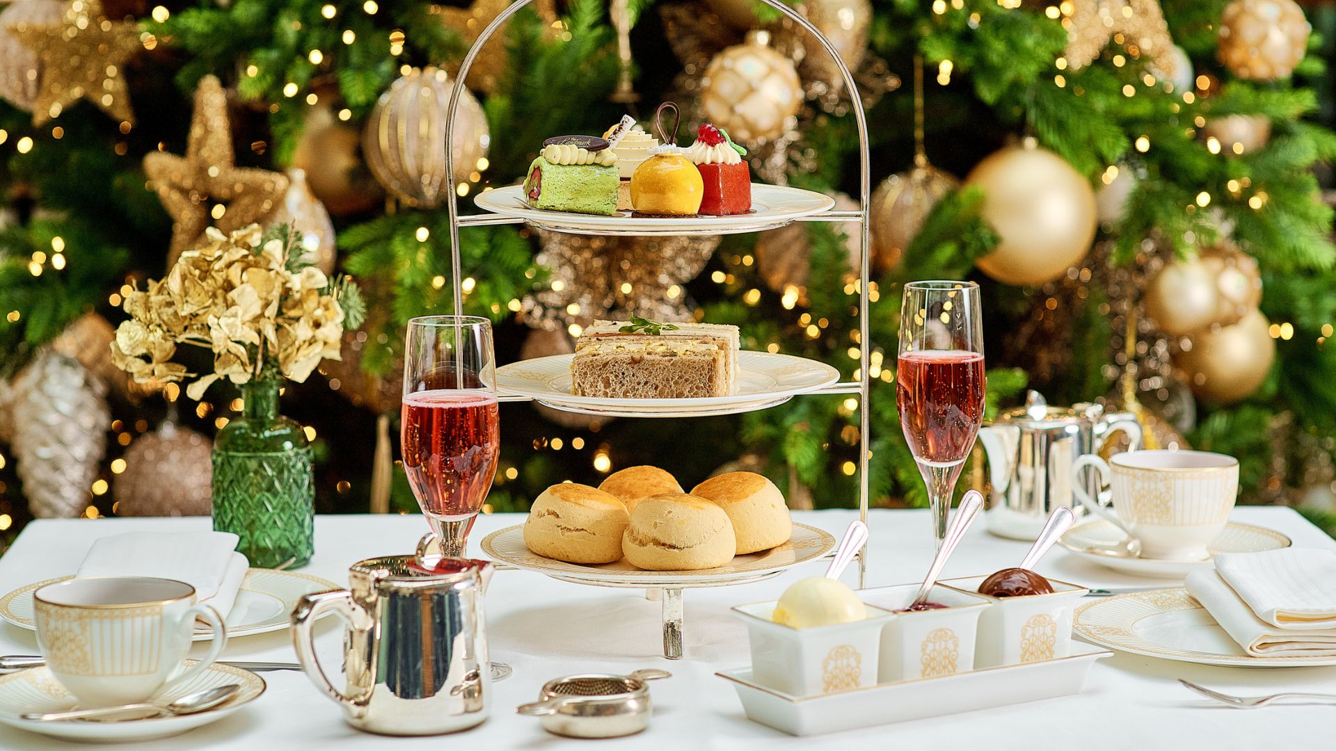 Festlicher Nachmittagstee im Winter Garden Restaurant im The Landmark London. Bild mit Genehmigung von Roche Communications. Festlicher Nachmittagstee mit einer dreistöckigen Etagere mit verschiedenen Backwaren, Sandwiches und Scones, dazu zwei Gläser Rosé-Sekt. Der Tisch ist mit edlem Porzellan, einer silbernen Teekanne und Gewürzen gedeckt, im Hintergrund steht ein geschmückter Weihnachtsbaum mit goldenen Ornamenten und funkelnden Lichtern.