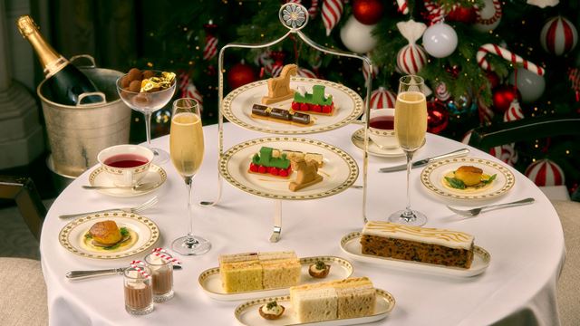 a large white table spread with an afternoon tea stand with sweet and savoury festive treats, plates of finger sandwiches on the table and cups of tea. A bottle of champagne sits in a bucket besides the table, and there is a decorated Christmas tree in the background.