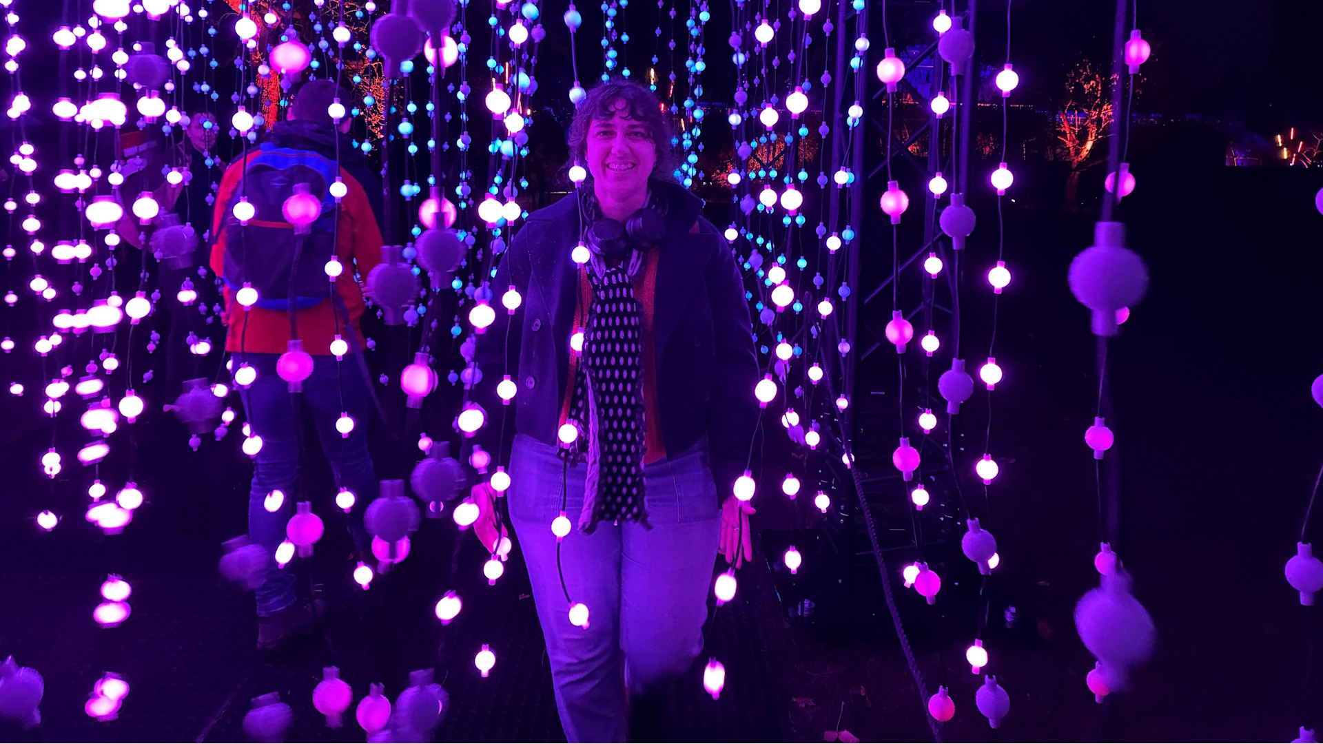 Become immersed in interactive light installations at Christmas at Kew. Photo credit: Jonny Payne © London & Partners. A woman walks between streams of vertical lights, illuminated in pinks, purples and whites, as part of the Christmas at Kew light trail.