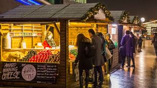 Des personnes se tiennent debout et parcourent les stands du marché de Noël dont un étal vendant du fromage.