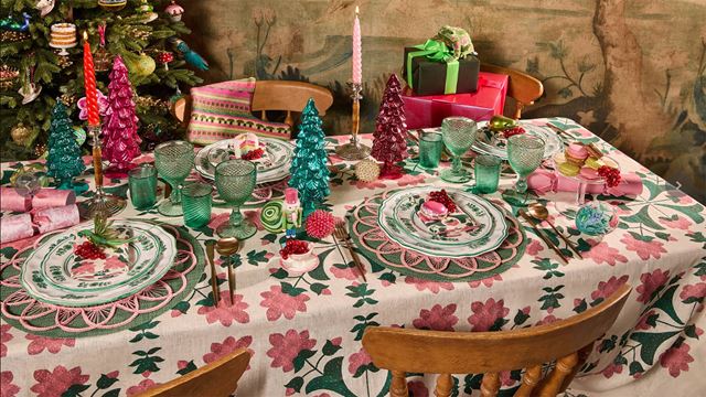 Une table festive est décorée dans des tons verts et roses avec des sets de table assortis, des bougies et des verres, des cadeaux de Noël et un sapin de Noël en arrière-plan.