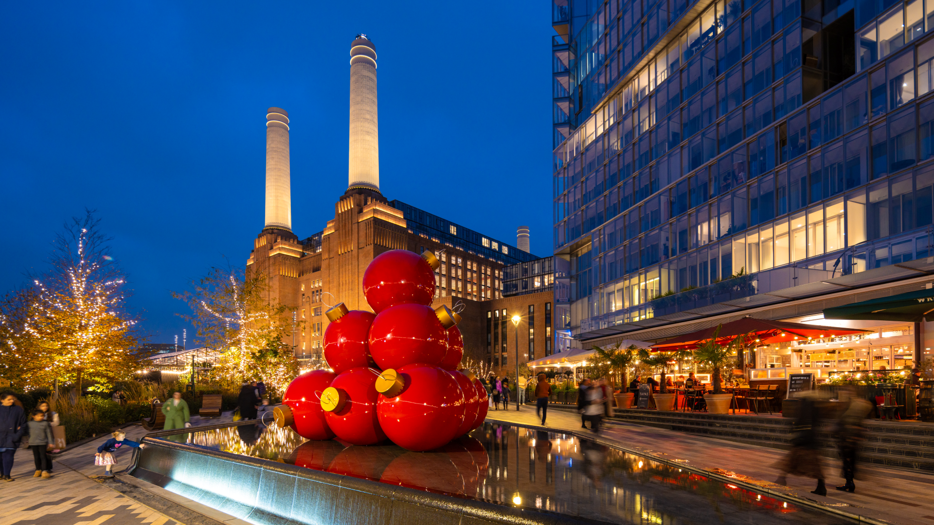 Sehen Sie, wie sich Battersea Power Station in der Weihnachtszeit verwandelt. Bildnachweis: Charlie Round-Turner. Bild mit Genehmigung von Battersea Power Station. Ein Stapel riesiger roter Christbaumkugeln steht vor der Battersea Power Station, während glühende Lichter und Weihnachtsbäume die Gegend erhellen