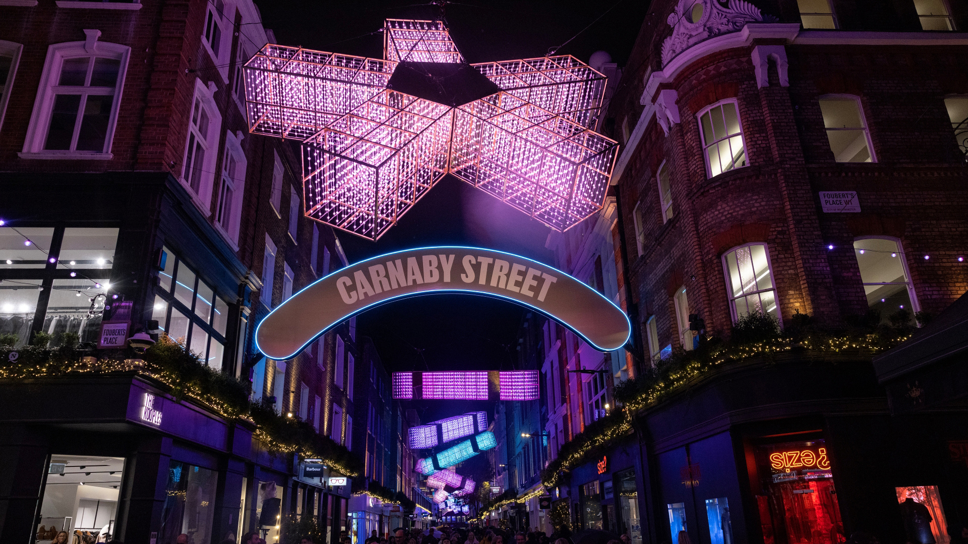 A photo of the 2025 Carnaby Christmas lights including abstract snowflake and cracker displays shining above the street in vibrant colours of orange, blue and pink