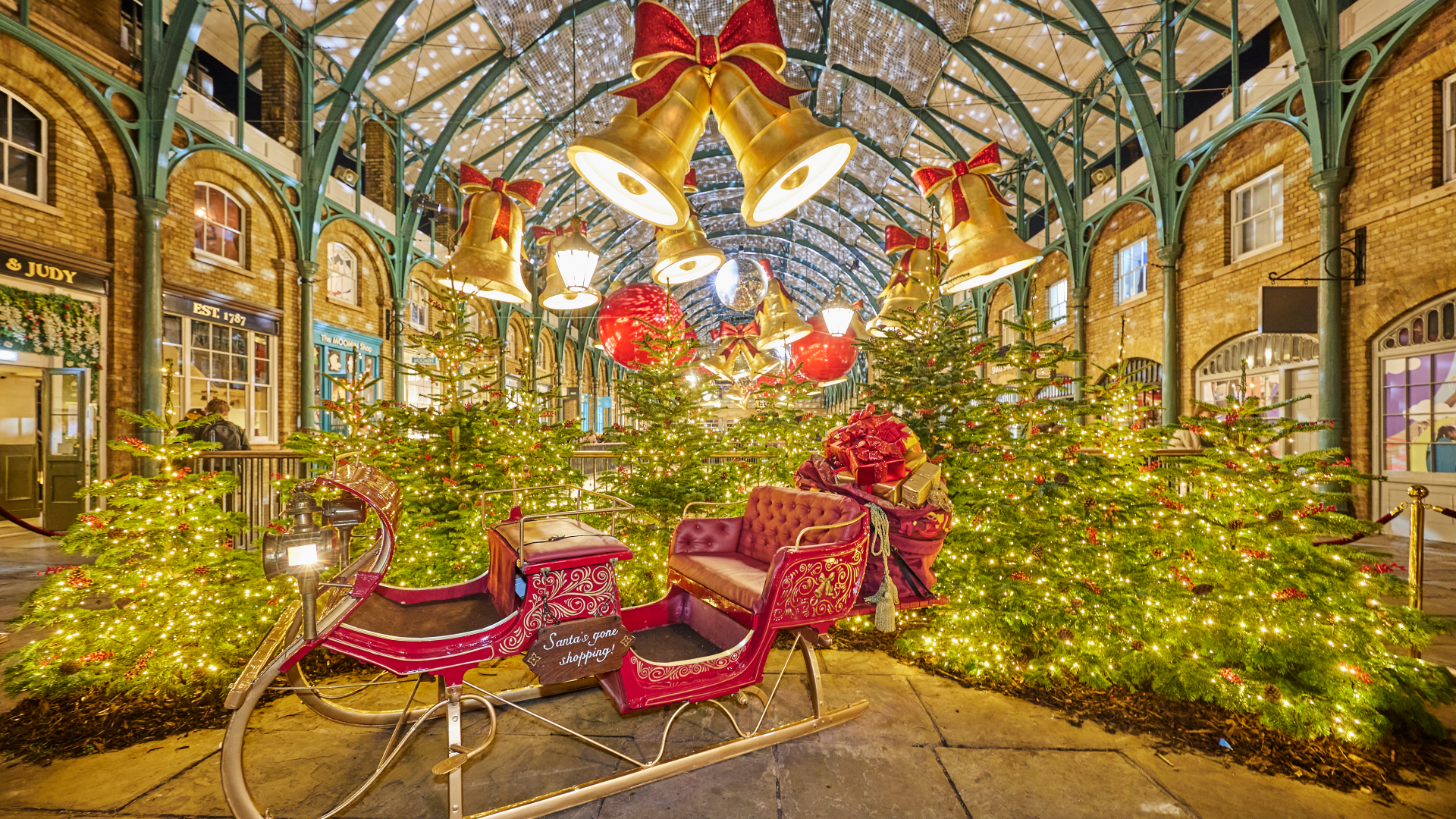 A red sleigh with gold details in the Covent Garden market surrounded by Christmas trees with twinkling lights with giant gold bells with bows hanging above