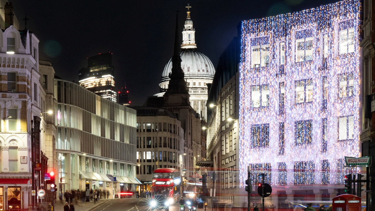 A photo of Ludgate Hill at night during winter with a building front covered in twinkling Christmas lights, St. Paul's Cathedral in the background lit up and red buses and cars on the street in the foreground 