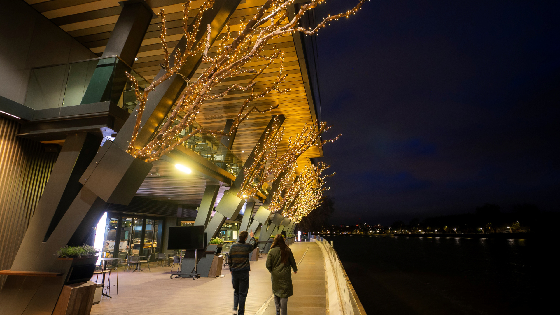 A photo of the riverside of Fulham Pier with giant branches wrapped with warm twinkling lights hanging from the side of the building as two people walk past
